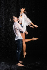 Wall Mural - happy boyfriend lifting smiling girlfriend in wet clothes in rain drops on black background