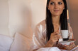© Lukas - A young beautiful female, wearing a skin-colored nightdress is sitting in a bed and holding a cup of coffee in her hands. Concept of a relaxed morning in bed.