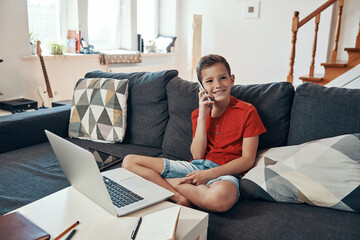 Wall Mural - Happy boy talking on the phone and looking at camera while homeschooling in the living room