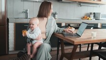 A Positive Mom Sitting At A Laptop And Holding Her Little Baby With A Pacifier In Her Hands In The Kitchen