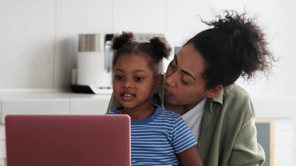 Wall Mural - A smiling african mother saying something in the ear of her little daughter and holding her in her arms while sitting in the kitchen