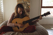© Wavebreak Media - Happy caucasian woman sitting on beanbag playing acoustic guitar in sunny cottage living room