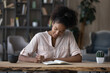 © fizkes - Smiling young mixed race african business woman sitting at table, writing notes in paper planner, managing working issues or making to-do list, planning workday or assignments at modern home office.