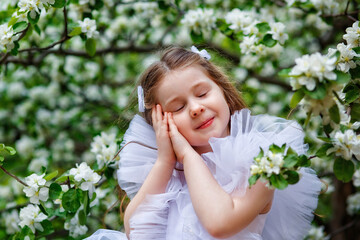 The girl closed her eyes dreamily and folded her hands over her face. A child plays under a blooming apple tree with white flowers. Summer outdoor activities in the spring garden