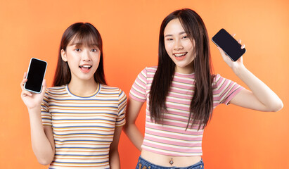 Wall Mural - Two beautiful young asian girls are using mobile phones on orange background