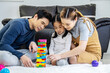 © TeTe Song - Happy asian family father and mother with little asian girl smiling playing with building tower from wooden blocks, taking tiles in turn from underneath until it falls, in moments good time at home