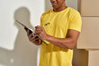 © Svitlana - Cropped shot of professional male mover in yellow t shirt making notes, holding clipboard, standing near a stack of cardboard boxes while doing home relocation