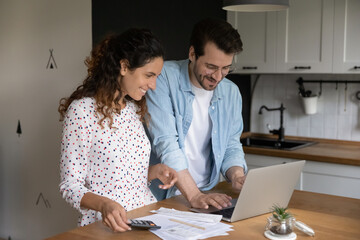 Happy bonding young married family couple using e-banking computer application, calculating domestic expenditures, planning future investment or vacation, paying utility bills or taxes online.