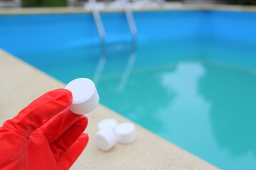 Wall Mural - White mini-tablet of chlorine in the hand of the staff for disinfection of swimming pools. The beginning of the swimming season is hot summer. Rubber protective red glove. Water purification.