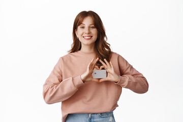 Wall Mural - Portrait of smiling redhead woman shows credit card, recommends bank, paying contactless, standing in casual clothes against white background