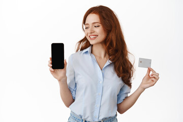 Smiling redhead businesswoman showing empty phone screen and credit card. Office lady looking satisfied at smartphone, advertising bank or store, standing against white background