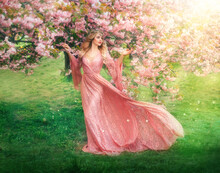 Fantasy Happy Girl Elf Princess Walks In Spring Blooming Garden. Pink Flowers Sakura Tree Green Grass Summer Nature. Long Lace Dress Wide Sleeves Flies In Wind Motion. Blonde Woman Queen. Vintage Gown