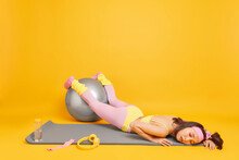 Workout Sport And Tiredness Concept. Exhausted Young Asian Woman Keeps Hands On Fitness Ball Feels Very Tired After Weary Training Dressed In Activewear Lies On Karemat Against Yellow Background
