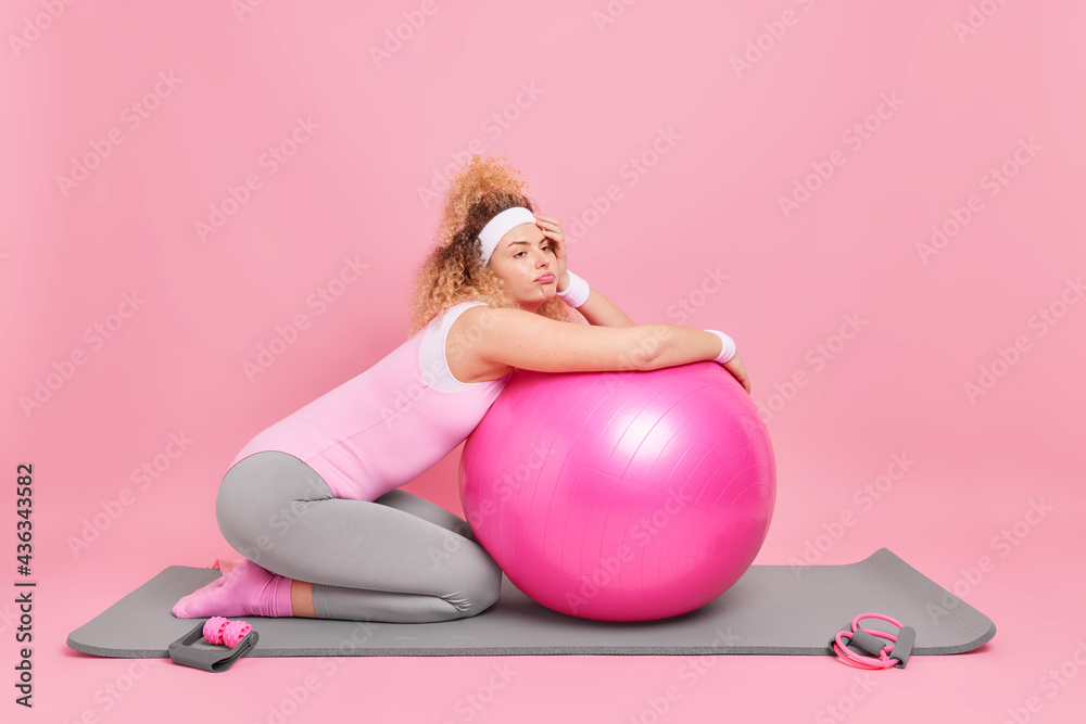 Displeased bored woman leans at fitness ball rests after exercise session dressed in activewear takes break from workout poses on karemat isolated over pink background. Health care pilates practice - obrazy, fototapety, plakaty 
