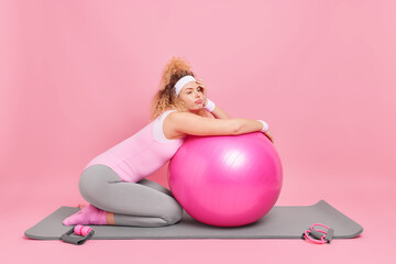 Displeased bored woman leans at fitness ball rests after exercise session dressed in activewear takes break from workout poses on karemat isolated over pink background. Health care pilates practice