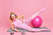 Cheerful curly woman with slim figure exercises on fitness mat raises legs leads active lifestyle poses against pink background sport equipment around. Pilates exercising. Athletic female has workout
