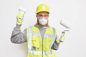 Construction and repair concept. Serious male builder uses painting tools busy doing house improvement wears protective respirator transparent glasses and uniform isolated over white background