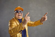 © Studio Romantic - Funny happy rich senior man in golden party jacket, baseball cap and bling gold chain necklace enjoying rap music. Old but youthful grandpa dancing and having fun isolated on gray studio background