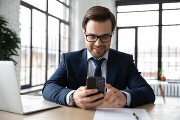 Wall Mural - happy businessman using online app on smartphone at office workplace. employee wearing suit and glas