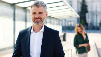 Camera approaching handsome happy Caucasian middle-aged bearded man in suit looking at camera and smiling waiting outdoor, traveling, man tourist, business trip, covid-19 pandemic, close up concept