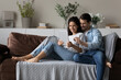 © fizkes - Smiling young multiethnic man and woman relax on couch look at tablet talk speak on video webcam call online. Happy multiracial couple rest at home use modern pad device shopping on internet.