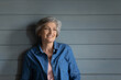 © fizkes - Mature attractive woman with grey short hair in denim shirt pose in studio standing leaned on blue wall, smile look into distance. Commercial services, anti-ageing product for older citizen ad concept