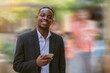 © arrowsmith2 - Confident african businessman using a smartphone and standing with smiling happy at the city