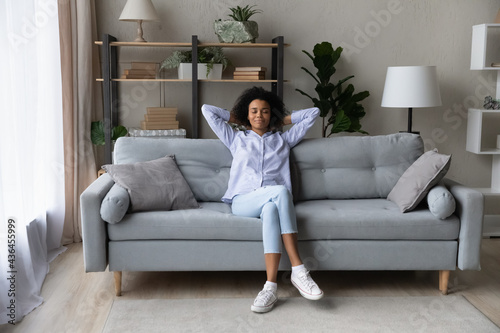 Happy calm young African American woman sit relax on sofa in living room take nap or sleep. Smiling millennial mixed race female renter rest on couch at home breathe fresh air. Rental concept.