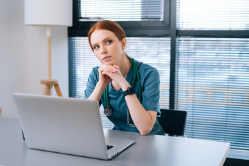 Portrait of thoughtful young female doctor in blue green medical uniform sitting at desk with laptop on background of window in hospital office of medic clinic, looking down.