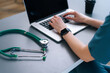 © dikushin - Back close-up view of hands of unrecognizable female practitioner wearing blue green medical uniform doctor using typing on laptop keyboard sitting at desk in modern office of medic clinic.