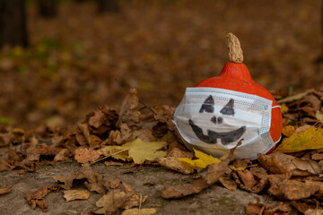 Wall Mural - Small orange pumpkin in medical mask with drawn face lies on fallen leaves in autumn forest. Selective focus. Copy space for your text. Theme of Halloween during coronavirus pandemic.