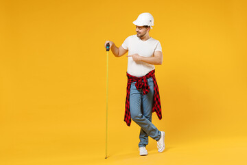 Full length young fun employee handyman man in protective helmet pointing index finger on measurement tape isolated on yellow background Instrument accessory renovation apartment room Repair concept.