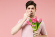 © ViDi Studio - Sick unhealthy ill allergic man has red eyes runny stuffy sore nose suffer from allergy symptoms hay fever hold blooming flower plant uses nasal drops isolated on pastel pink color background studio.