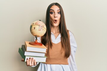 Wall Mural - Beautiful brunette young woman holding pile of books and vintage world ball scared and amazed with open mouth for surprise, disbelief face