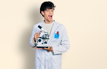 Sticker - Handsome hipster young man wearing lab coat holding microscope winking looking at the camera with sexy expression, cheerful and happy face.