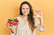 Young hispanic girl holding strawberries smiling and confident gesturing with hand doing small size sign with fingers looking and the camera. measure concept.