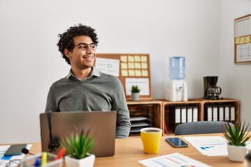 Poster - Young hispanic man wearing business style sitting on desk at office looking away to side with smile on face, natural expression. laughing confident.