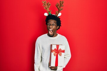 Sticker - Young african american man wearing deer christmas hat holding gift angry and mad screaming frustrated and furious, shouting with anger. rage and aggressive concept.