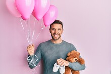Handsome Man With Beard Expecting A Baby Girl Holding Balloons, Shoes And Teddy Bear Smiling With A Happy And Cool Smile On Face. Showing Teeth.