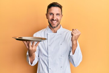 Sticker - Handsome man with beard wearing chef uniform holding silver tray screaming proud, celebrating victory and success very excited with raised arm