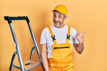 Poster - Handsome middle age man with grey hair holding ladder smiling with happy face looking and pointing to the side with thumb up.