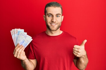 Poster - Young caucasian man holding swedish krona banknotes smiling happy and positive, thumb up doing excellent and approval sign