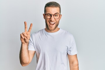 Sticker - Young caucasian man wearing casual clothes and glasses smiling with happy face winking at the camera doing victory sign. number two.