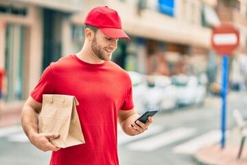 Canvas Print - Young caucasian deliveryman using smartphone and holding delivery paper bag at the city.