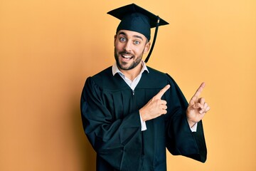 Wall Mural - Young hispanic man wearing graduation cap and ceremony robe smiling and looking at the camera pointing with two hands and fingers to the side.