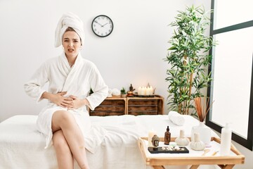 Poster - Young blonde woman wearing bathrobe at wellbeing spa with hand on stomach because indigestion, painful illness feeling unwell. ache concept.