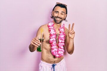 Young hispanic man wearing swimsuit and hawaiian lei drinking tropical cocktail smiling looking to the camera showing fingers doing victory sign. number two.