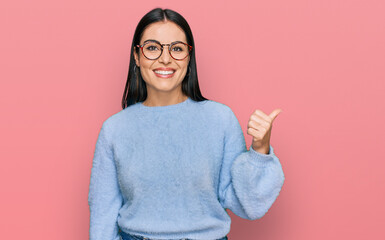 Wall Mural - Young hispanic woman wearing casual clothes and glasses smiling with happy face looking and pointing to the side with thumb up.