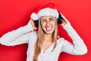 Sticker - Beautiful hispanic woman wearing christmas hat and headphones winking looking at the camera with sexy expression, cheerful and happy face.