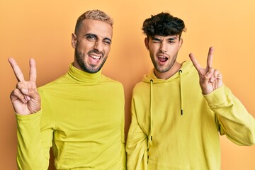 Wall Mural - Homosexual gay couple standing together wearing yellow clothes smiling with happy face winking at the camera doing victory sign. number two.